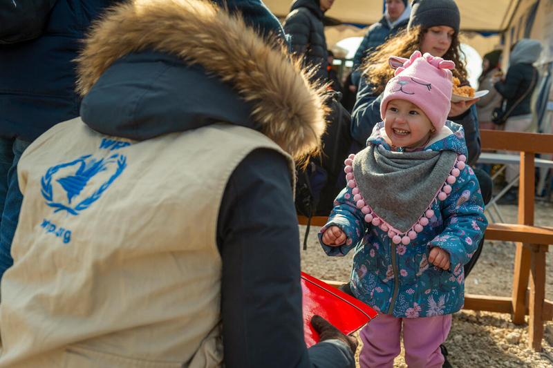 Una bambina sorridente, con un cappello e una sciarpa rosa sorride ad una operatrice del WFP inginocchiata davanti a leiFood distribution at the Ukraine-Moldova border reception center near the town of Palanca.