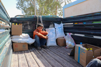 un ragazzo con una maglietta arancione e jeans blu si accovaccia sul retro di un camion in Siria, tra borse e scatole contenenti cibo del WFP