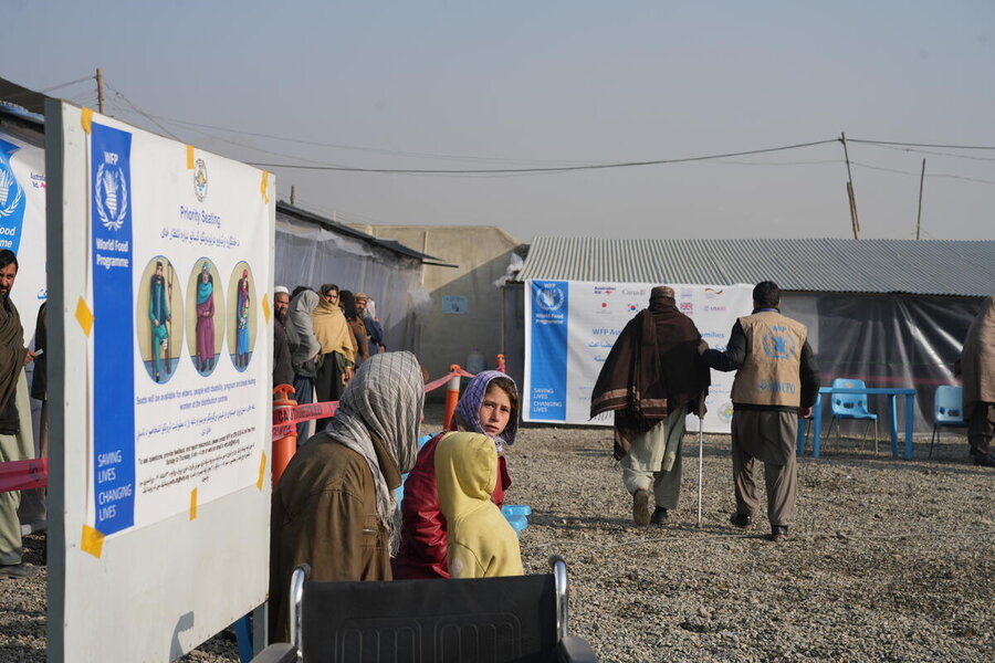 In an outdoor waiting area a girl looks into the camera as a person with disabilities is escorted away